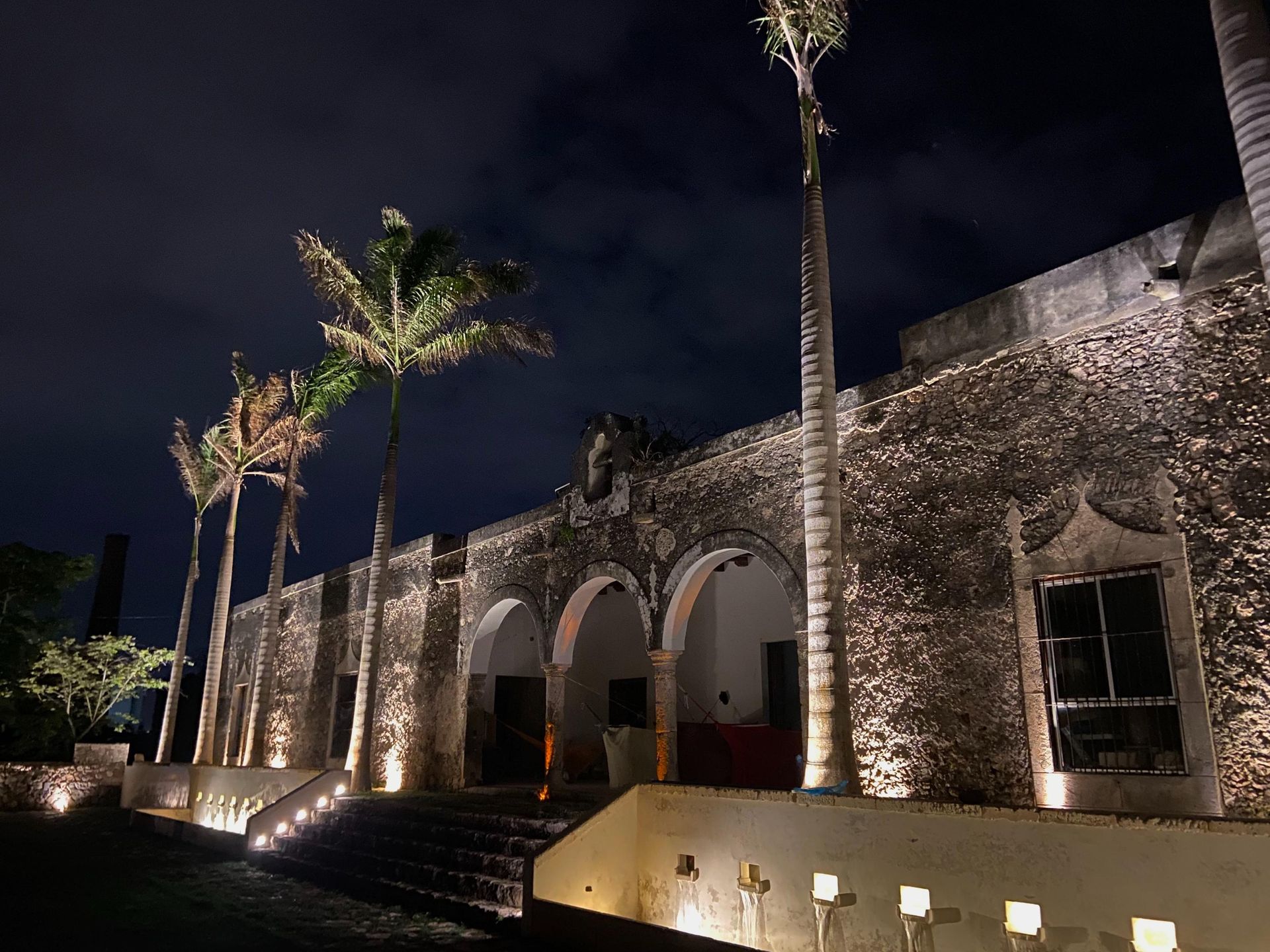 A stone building is lit up at night with palm trees in front of it.