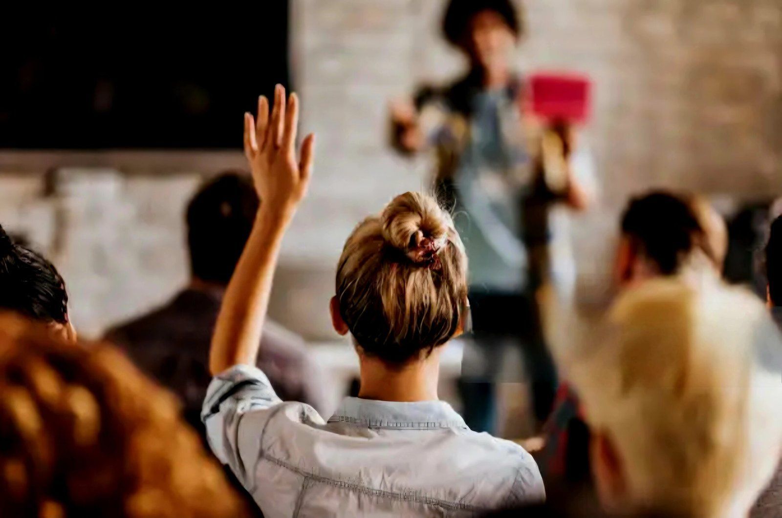Woman raising hand in a presentation, audience in foreground, presenter holding tablet in background.