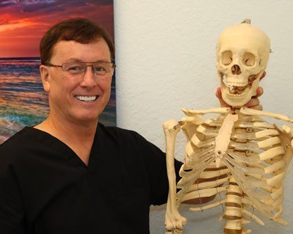 Man in black scrubs holds a skeleton. He smiles beside a beach scene.
