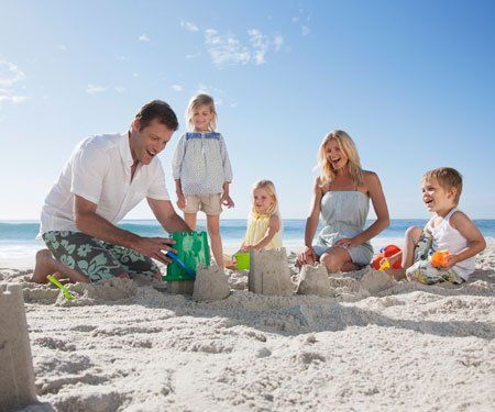 Family building sandcastles on a sunny beach, smiling, blue sky and ocean in background.