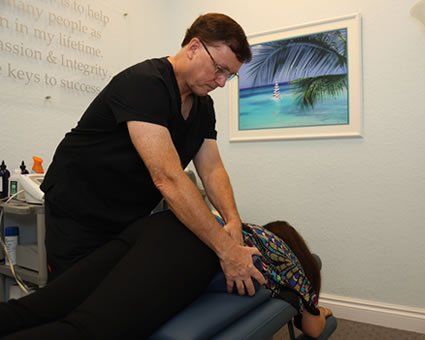 Chiropractor adjusting a patient's back on an examination table. White-walled room, neutral colors, small ocean picture.