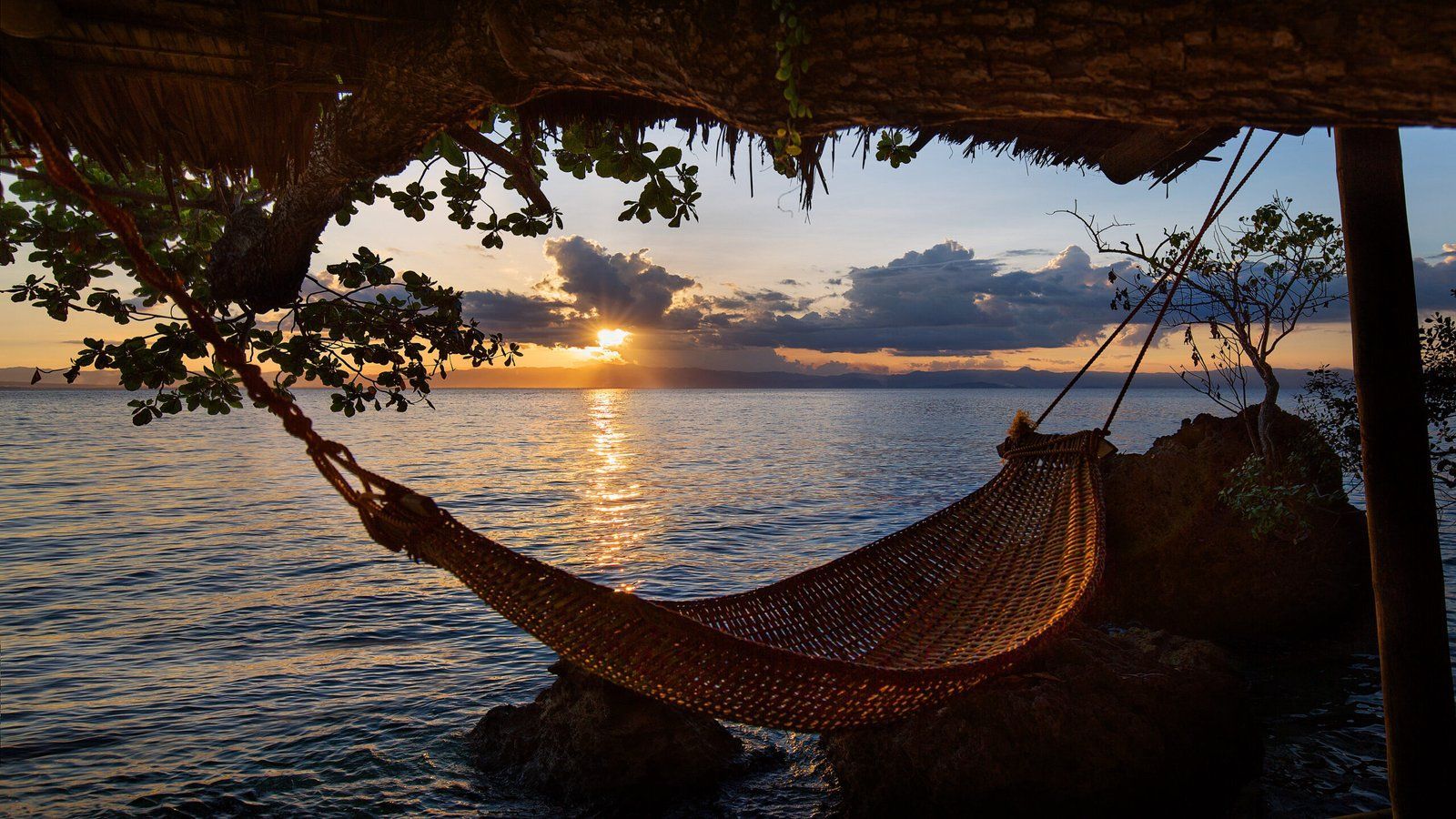Hammock strung over ocean at sunset. Sun reflects on water.