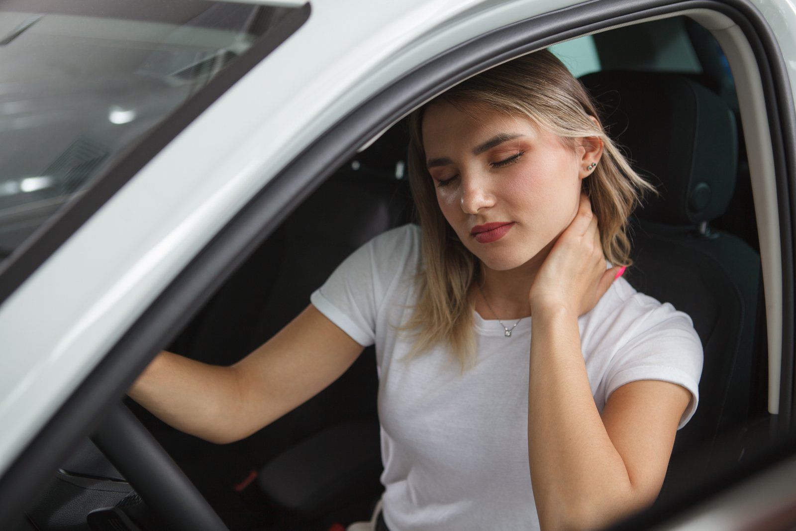 Woman in a white car, touching her neck, possibly in pain.