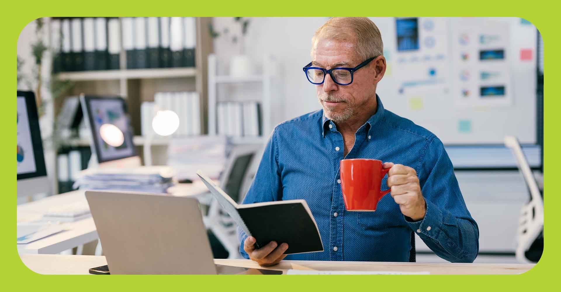 A business man in glasses and a collared shirt drinking coffee and looking at a book in an office