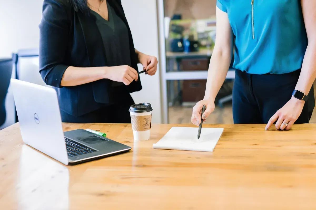 Two women are standing at a table with a laptop and a cup of coffee.