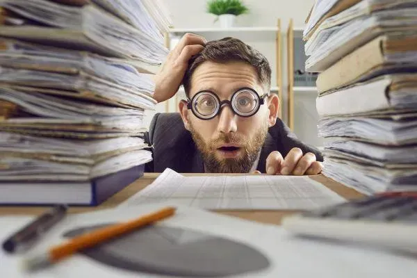 A man wearing glasses is sitting at a desk surrounded by stacks of papers.