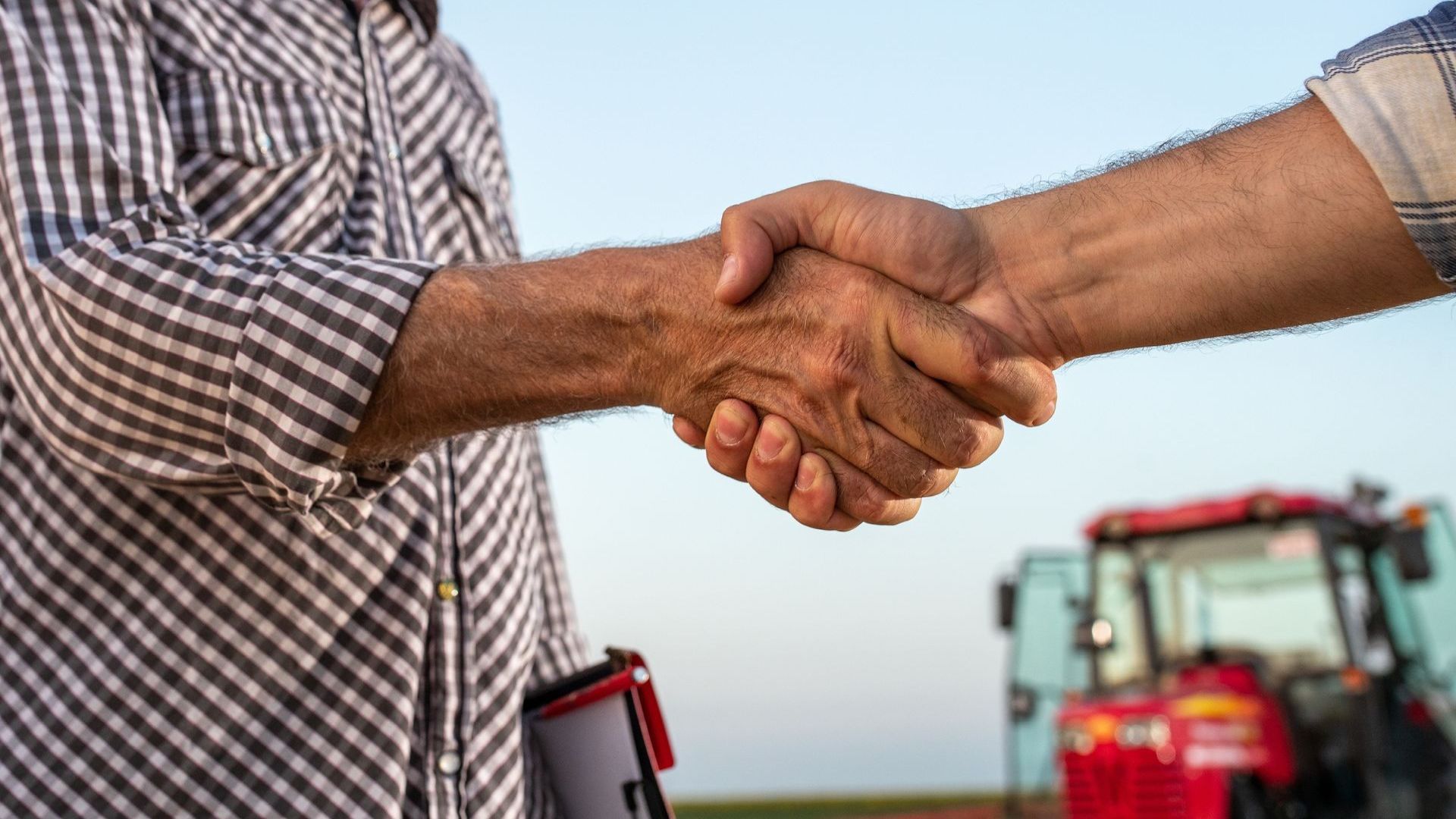 Two men are shaking hands in a field with a tractor in the background.