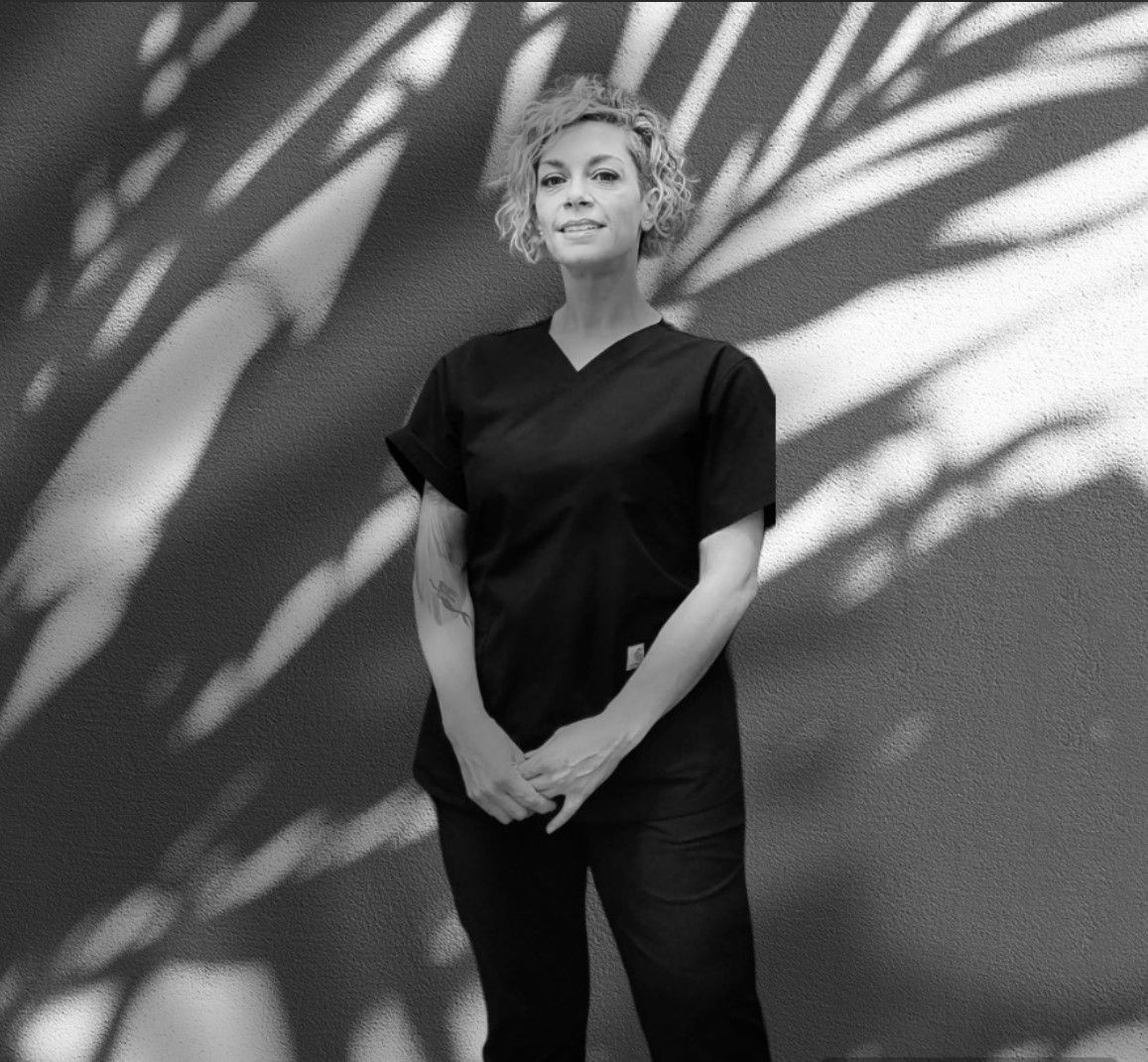 A black and white photo of a woman in scrubs standing in front of a wall.