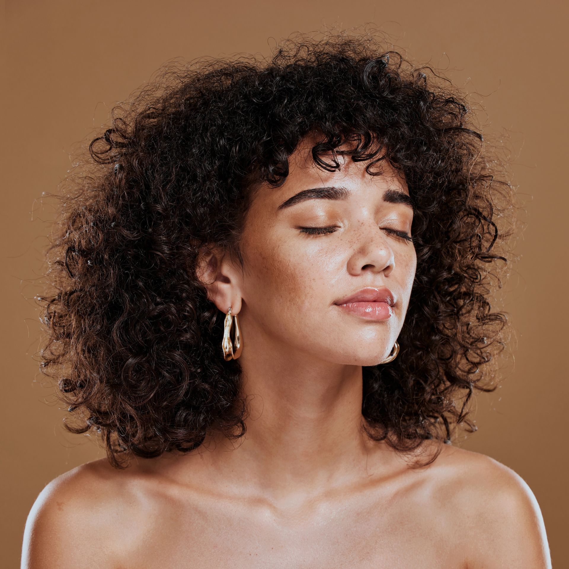 A woman with curly hair and hoop earrings is looking down with her eyes closed.