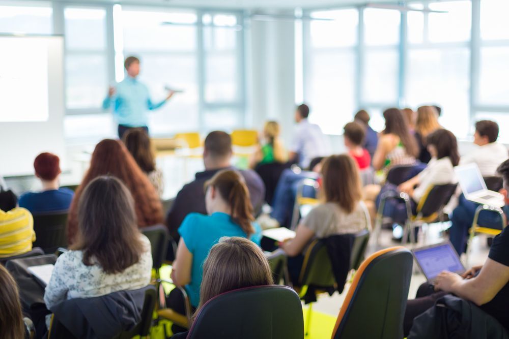 Audience in chairs listening to a presenter at a conference, blurred background.