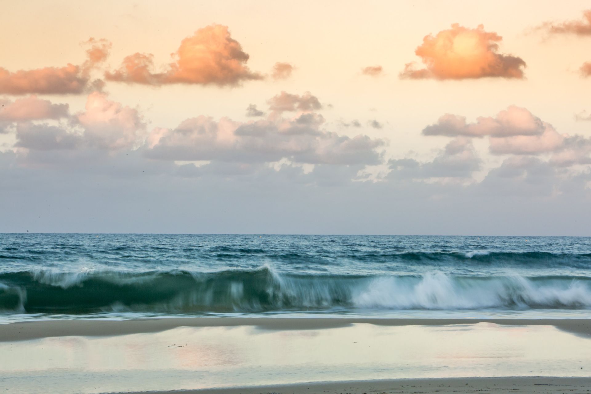 Ocean waves crashing on a sandy beach under a cloudy, pastel-coloured sky at dusk.