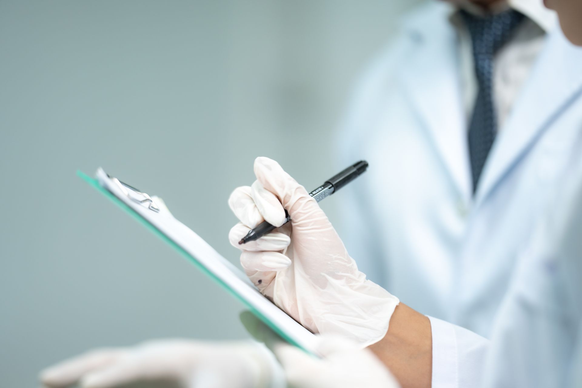 Person in lab coat and gloves writing on a clipboard, another person in the background.