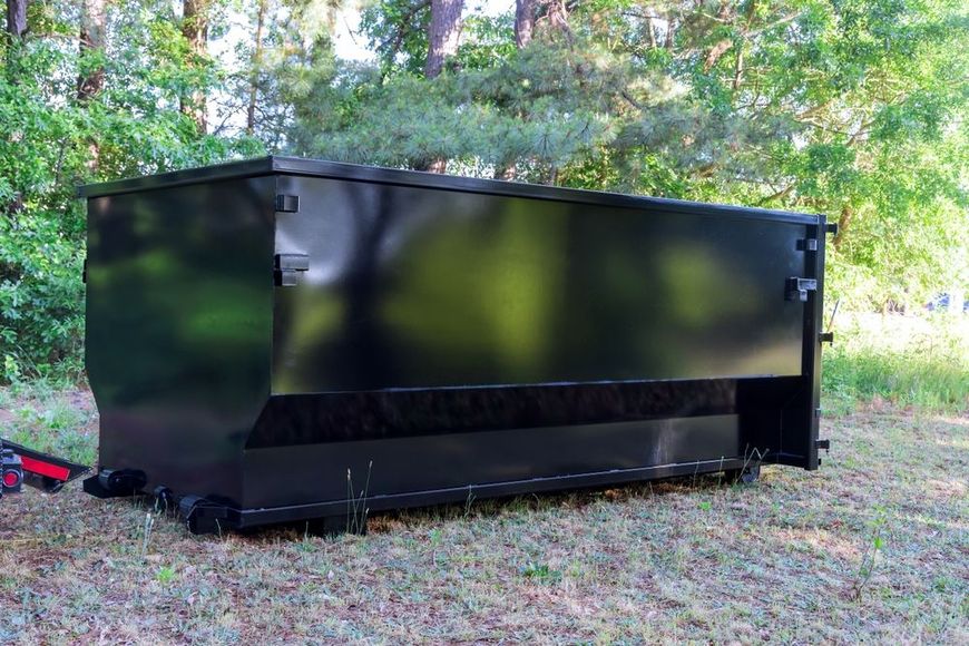 A black dumpster is sitting on top of a lush green field.