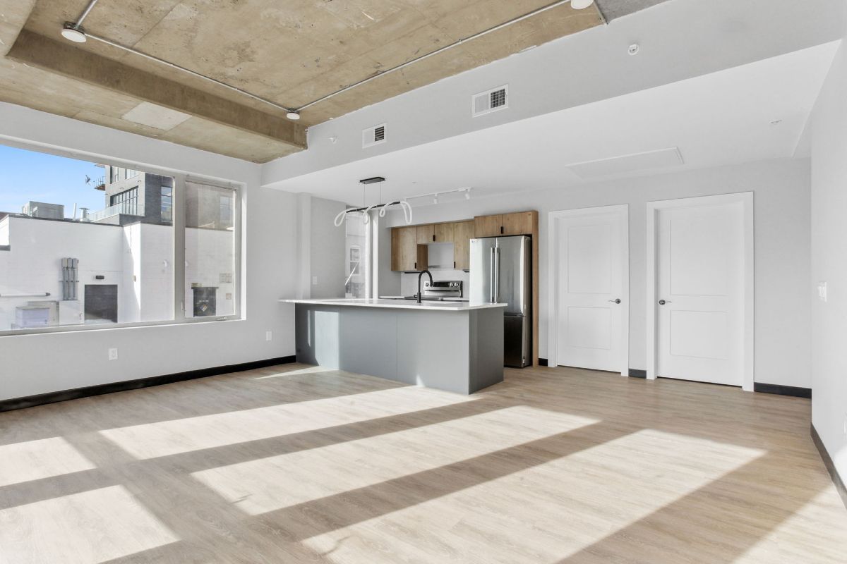 An empty living room with a kitchen and a large window.
