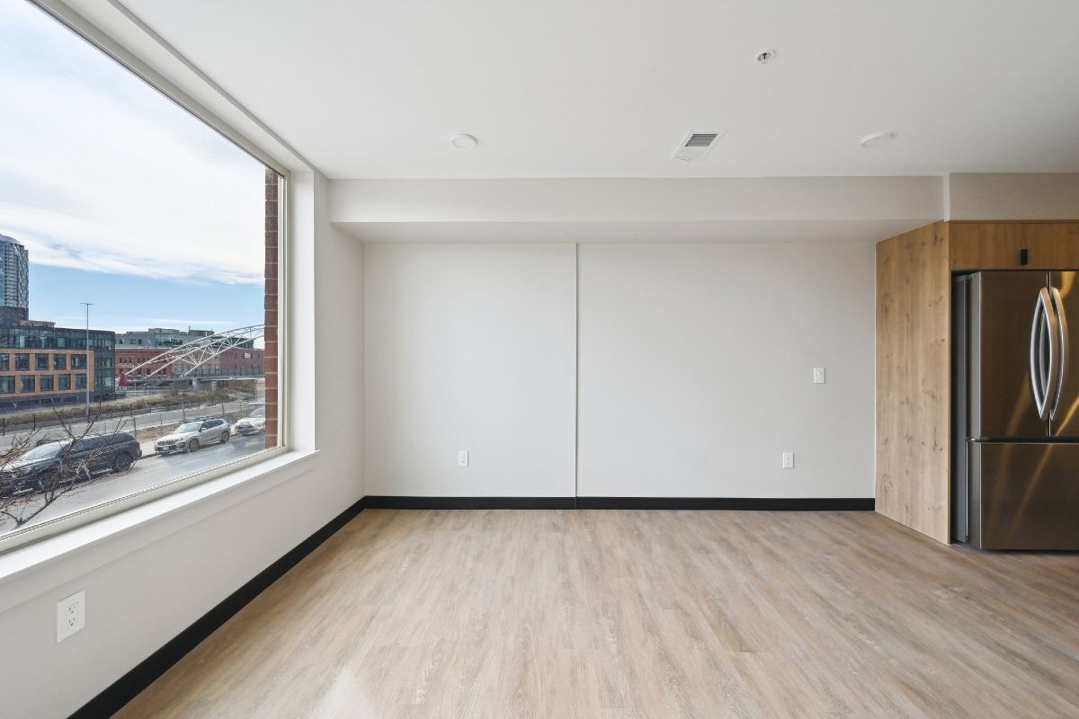 An empty living room with a large window and a refrigerator.