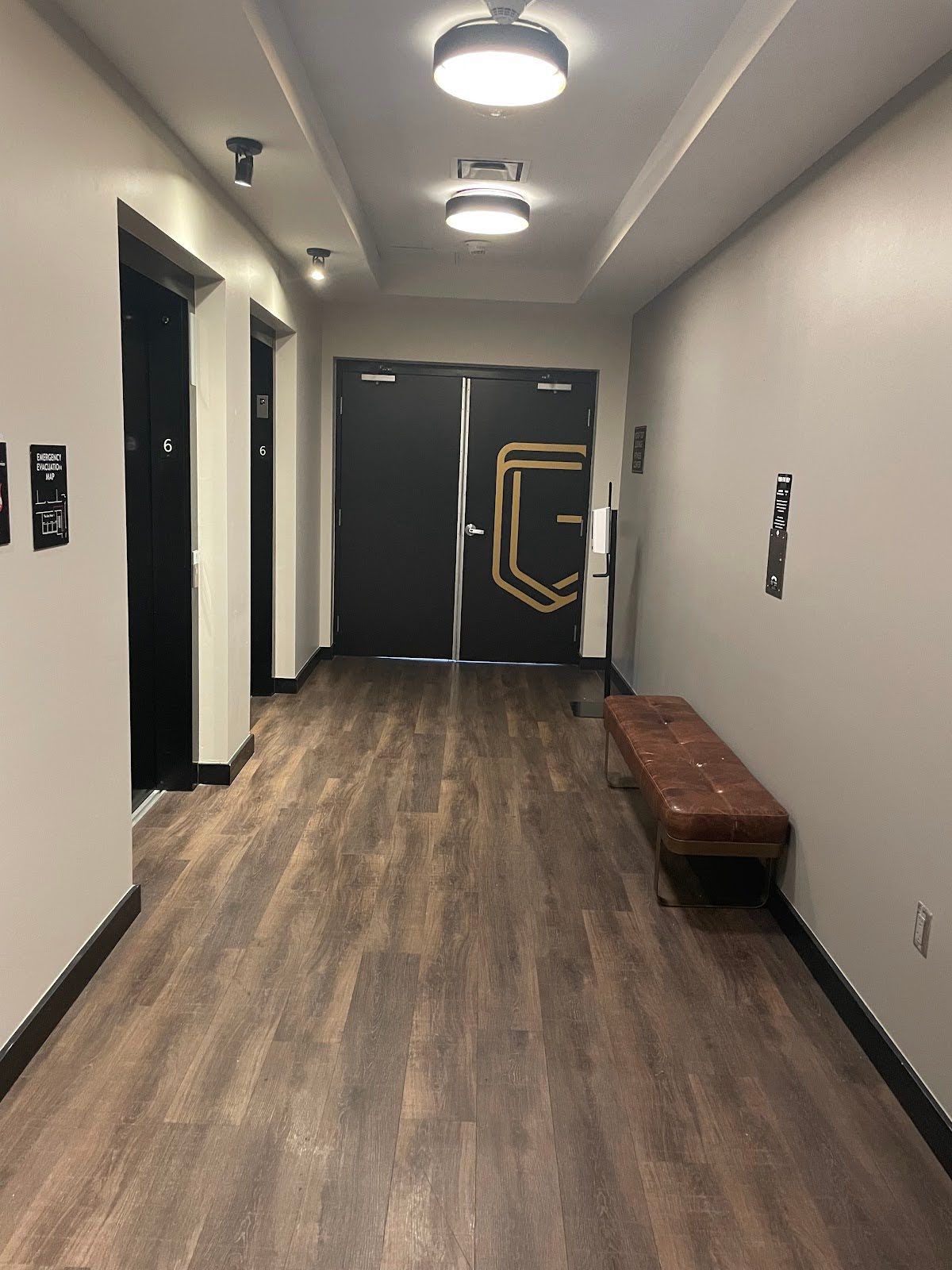 Hallway with elevators, brown wooden floor, gray walls, and a brown bench.