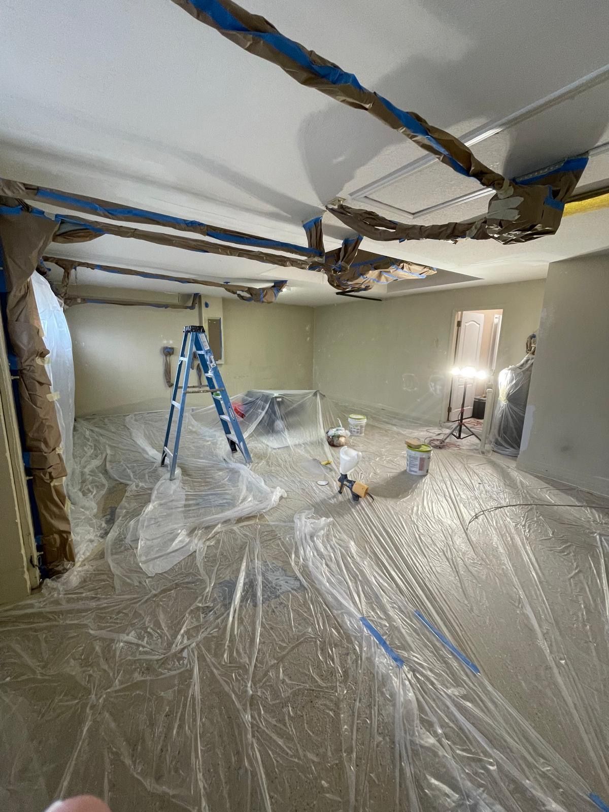 Room under renovation, covered in plastic, with blue ladder and masking tape on beams.
