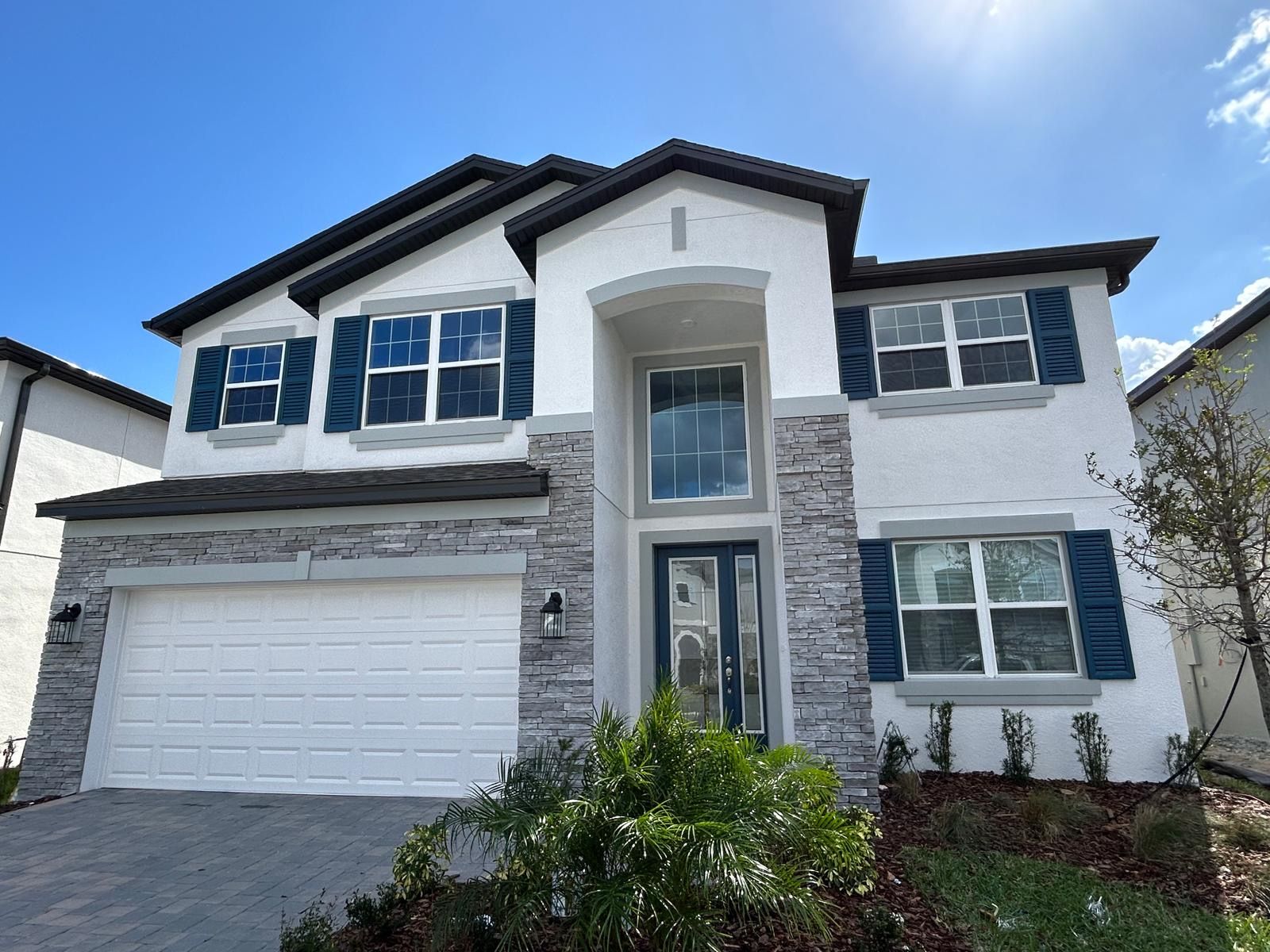 Two-story house with white stucco, stone accents, blue shutters, and a two-car garage under a blue sky.