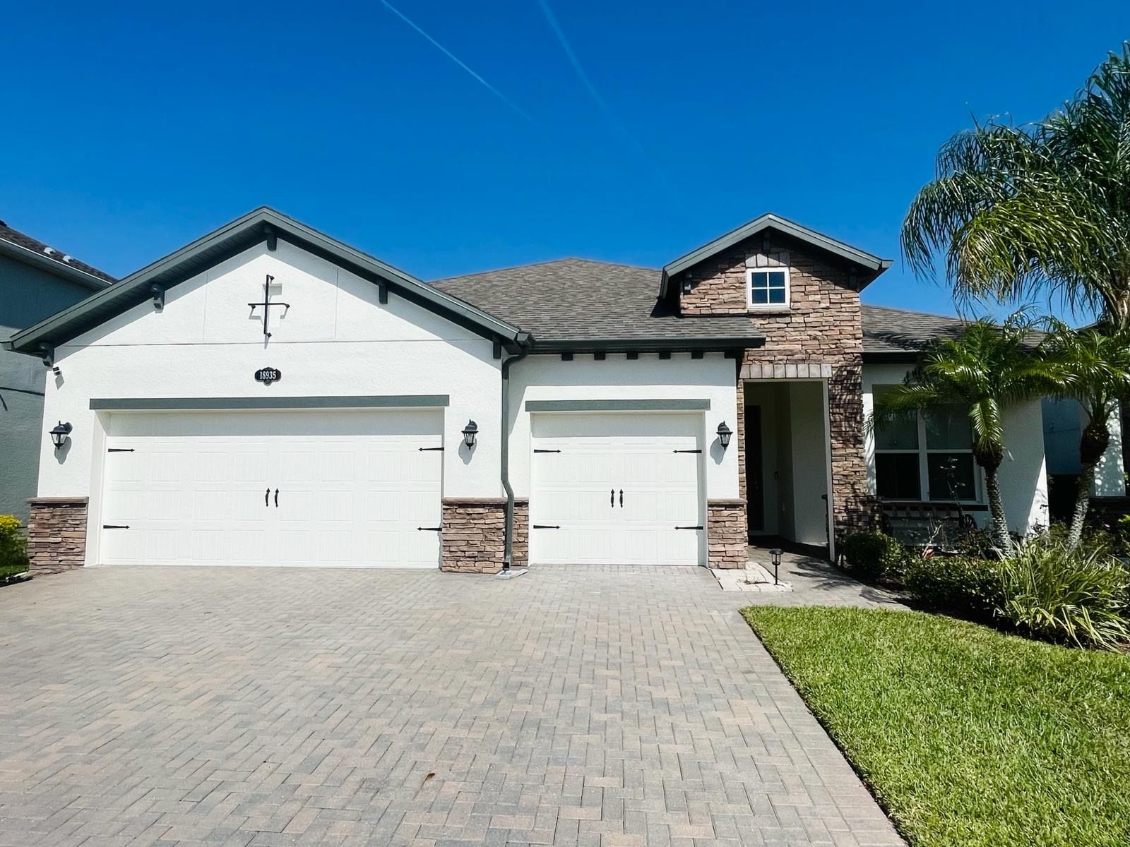 White house with two-car garage, brick accents, paved driveway, and palm tree under a blue sky.