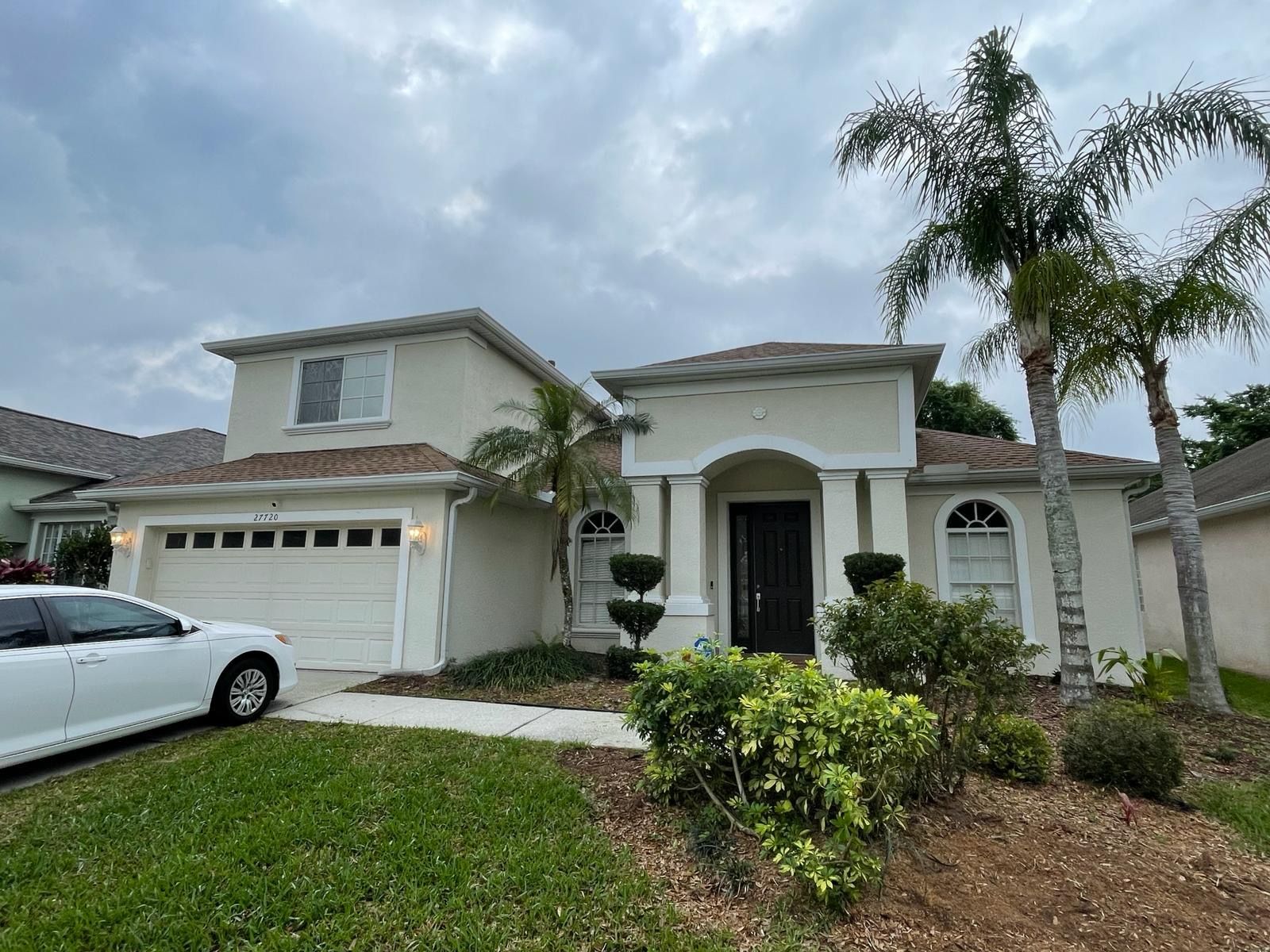 Beige two-story house with a white garage door, palm trees, and a white car parked in the driveway.