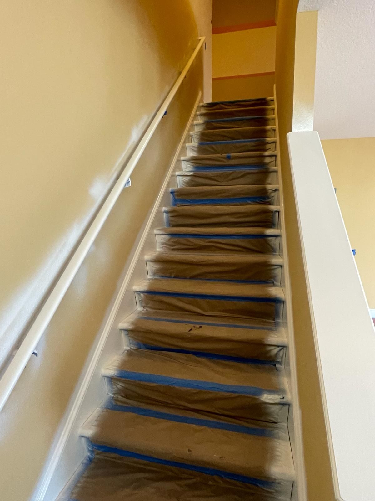 Staircase being painted. Steps covered with blue tape and plastic. Handrail along the left. Beige walls.