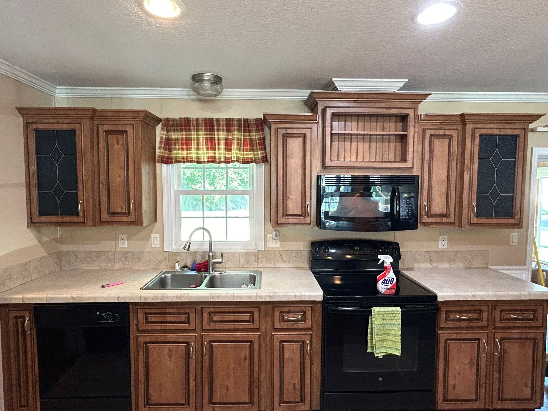 Kitchen with brown cabinets, black appliances, and a window with a plaid valance.