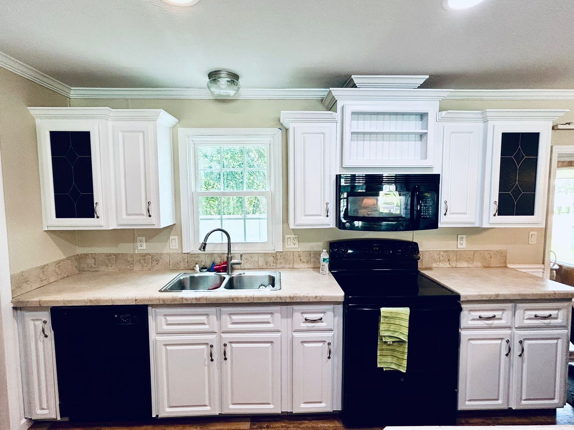 White kitchen cabinets and countertops with black appliances and sink under a window.