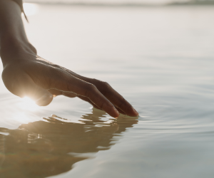 Picture of a hand touching the water with gentle ripples and a reflection of light below the thumb