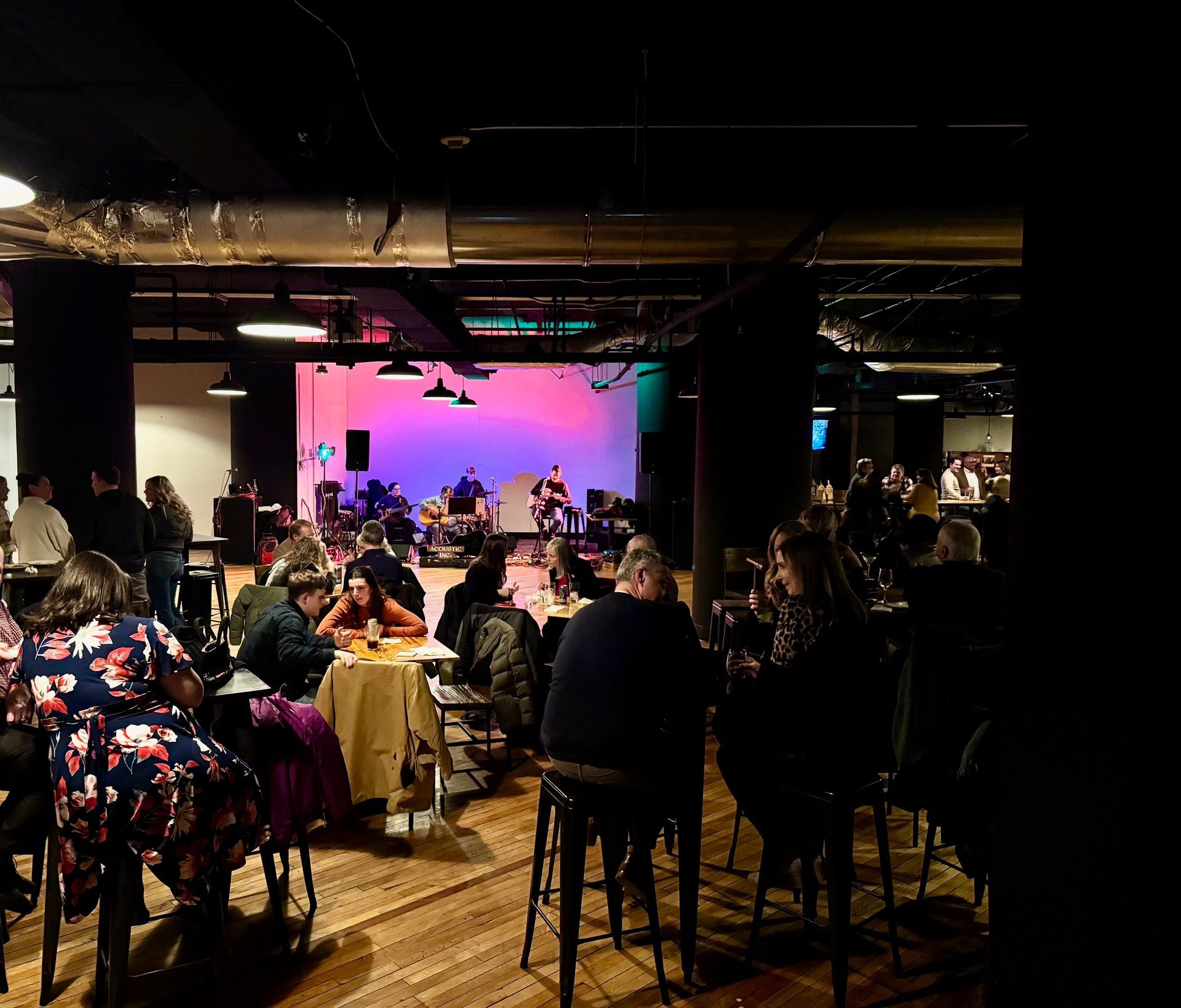 People seated at tables in a dimly lit venue, stage in background with band.