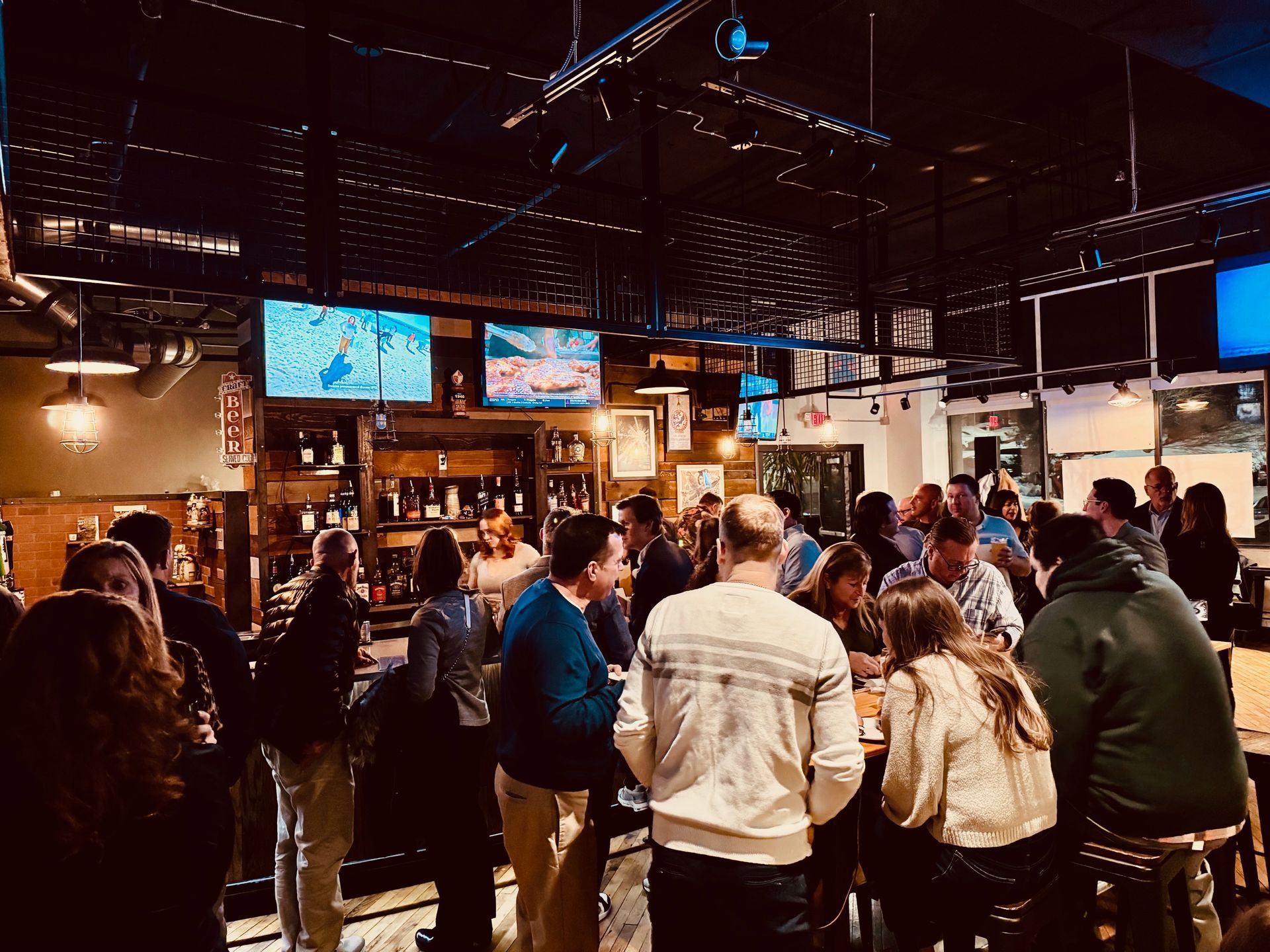 Crowd of people socializing in a dimly lit bar, watching TV screens.