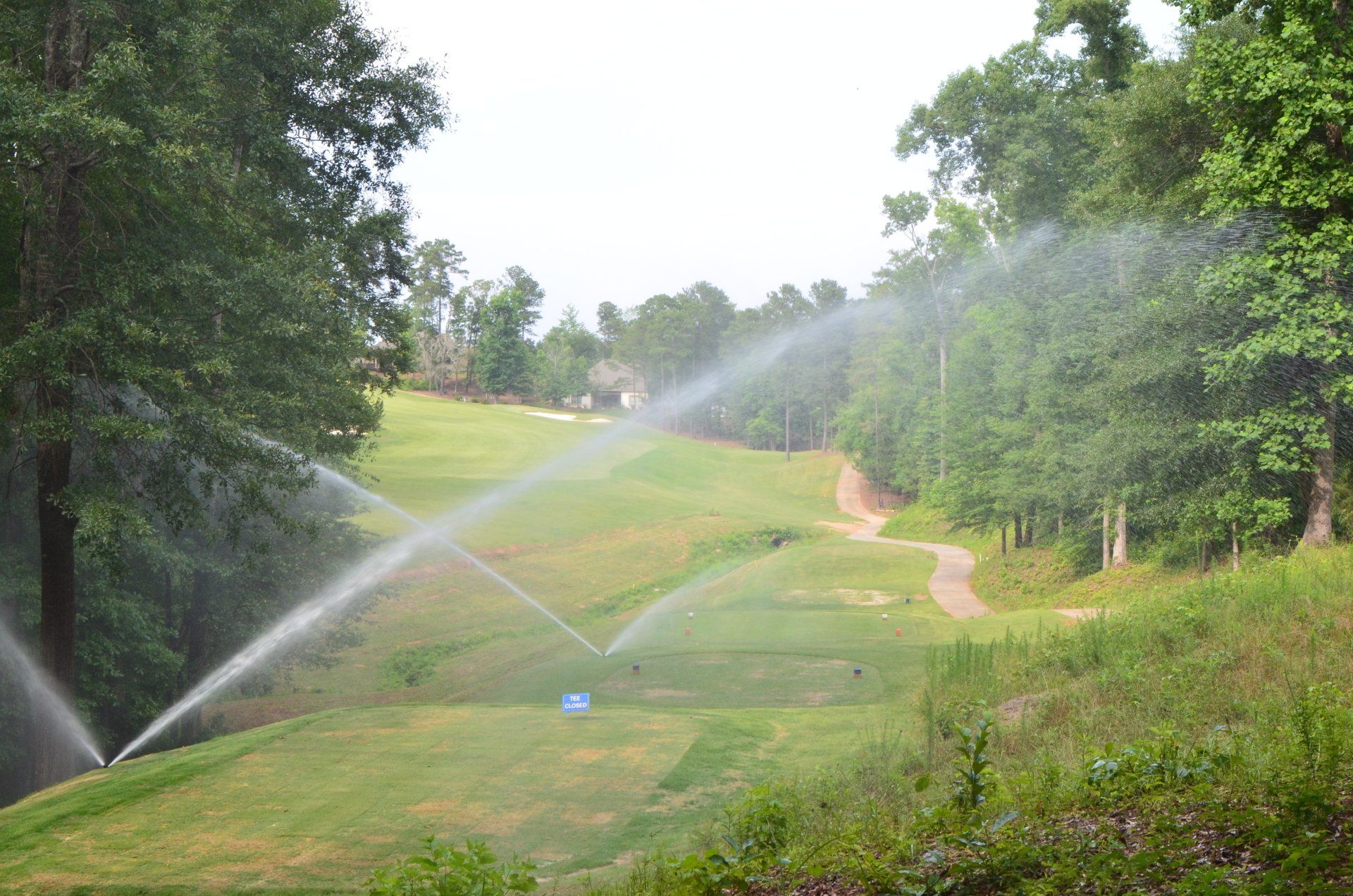 Two sprinklers are spraying water on a golf course surrounded by trees.