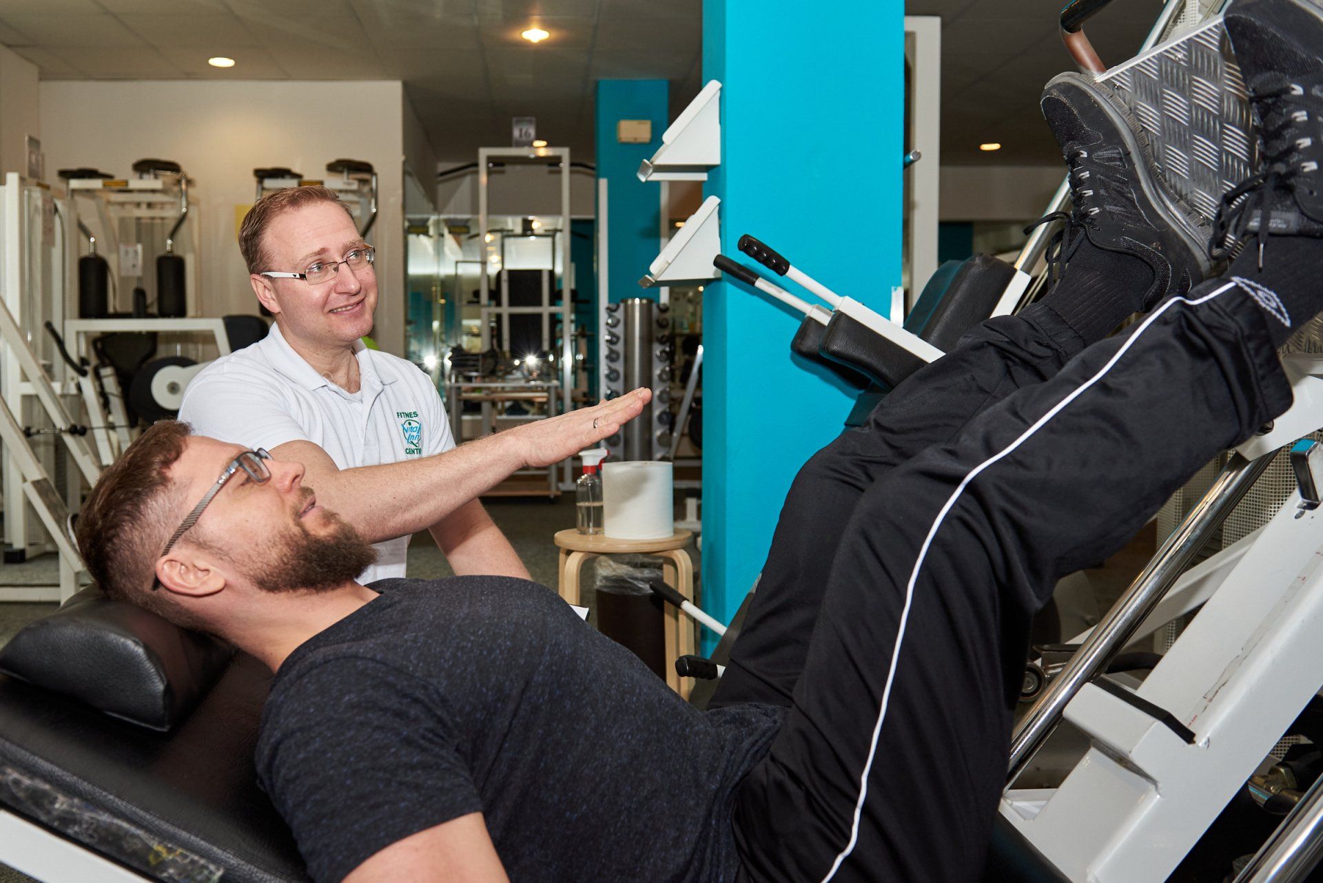 Ein Mann liegt in einer Turnhalle auf einer Bank, während ein Mann mit ihm spricht.