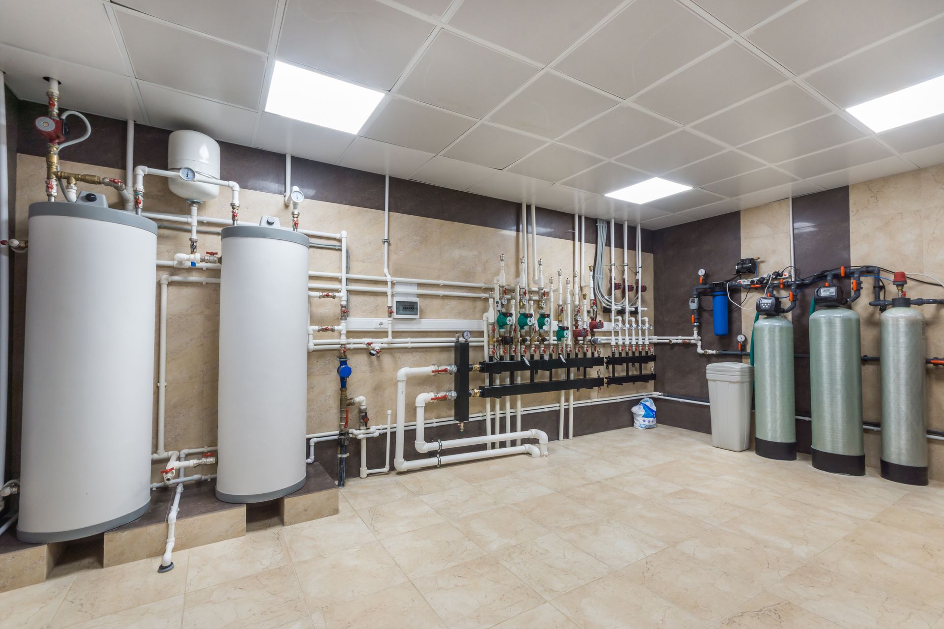 A mechanical room featuring two large white boilers on the left, pipes along the wall, and three tall water tanks.