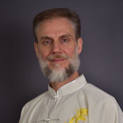 Un uomo con la barba indossa una camicia bianca e guarda la telecamera.