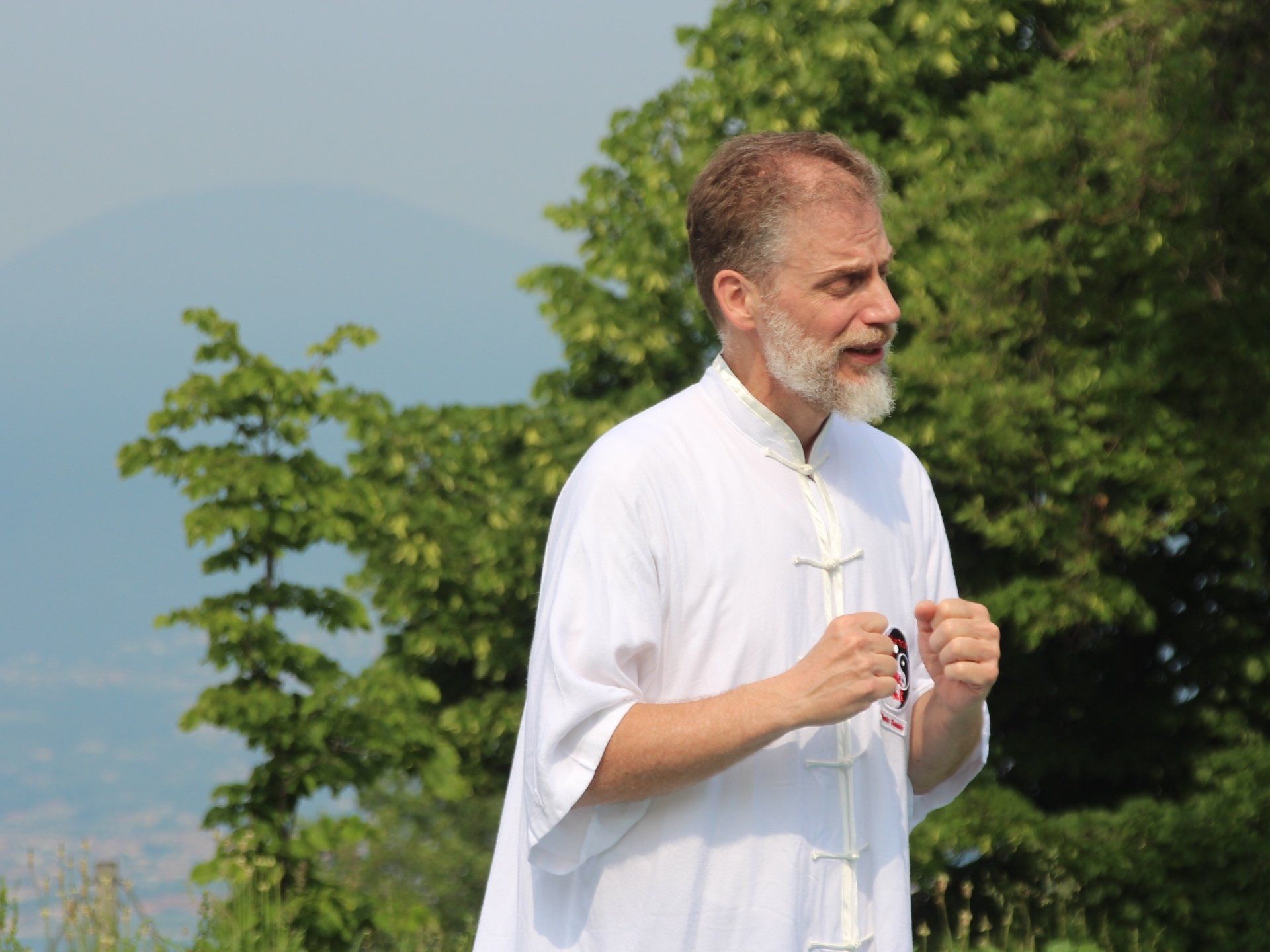 Un uomo con la barba indossa una camicia bianca e sta in piedi davanti agli alberi.