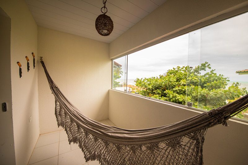 Uma rede de tecido bege está pendurada em uma varanda com paredes brancas, uma luminária pendente e uma grande janela com vista para árvores verdes.
