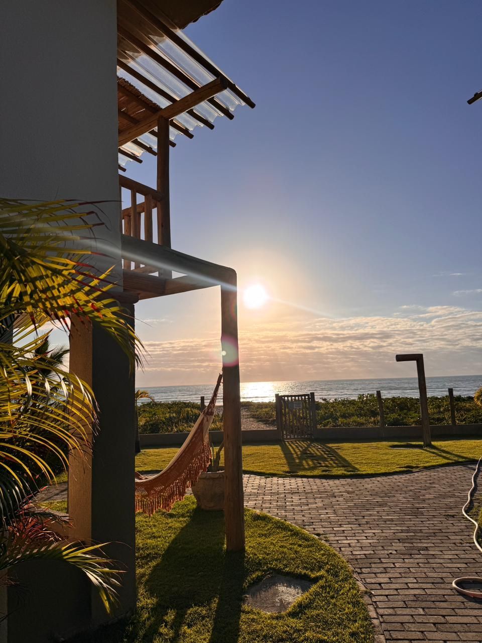 Um nascer do sol sobre o oceano visto de um pátio com uma rede e um caminho de tijolos, ao lado de uma casa de vários andares.