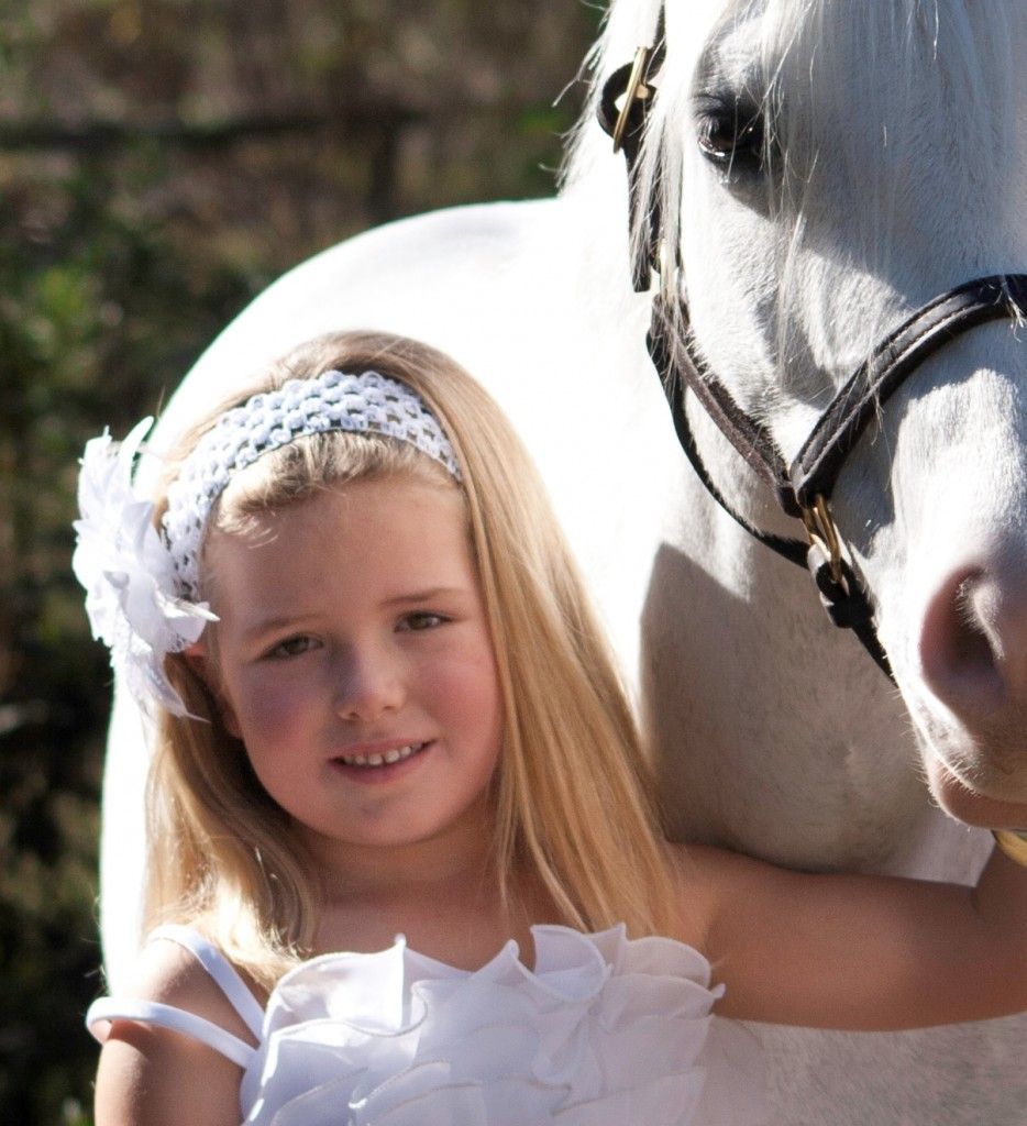 A little girl in a white dress stands next to a white horse