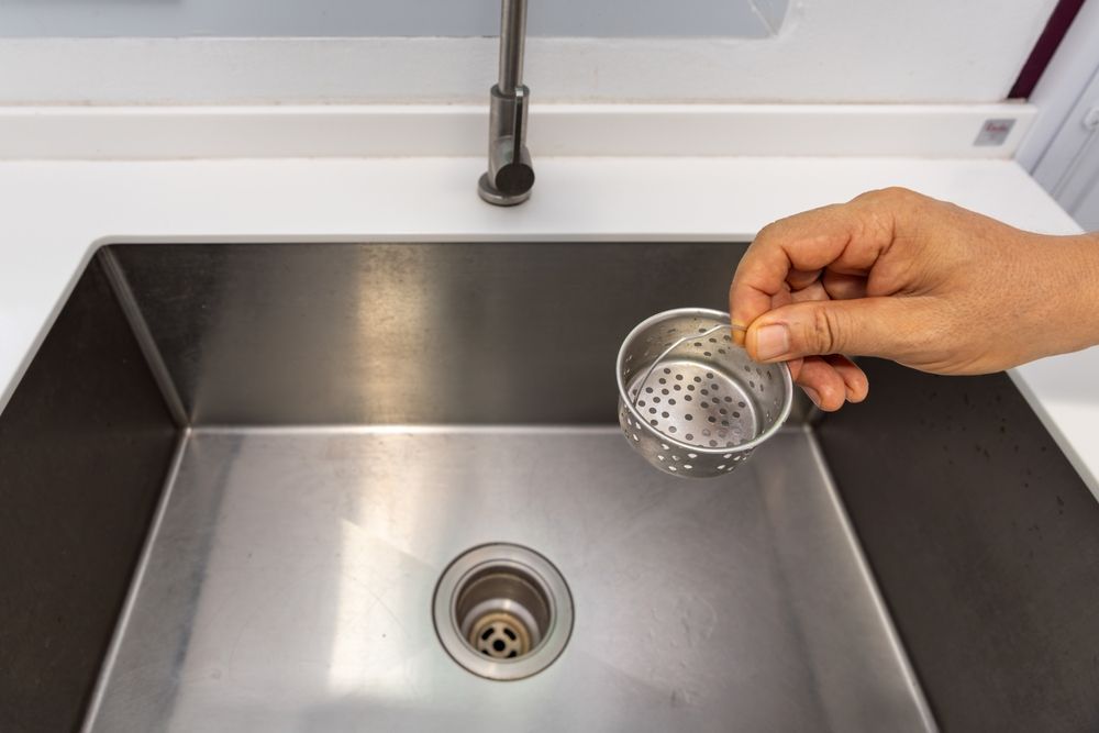 A person holds a strainer over a stainless steel sink with a faucet.