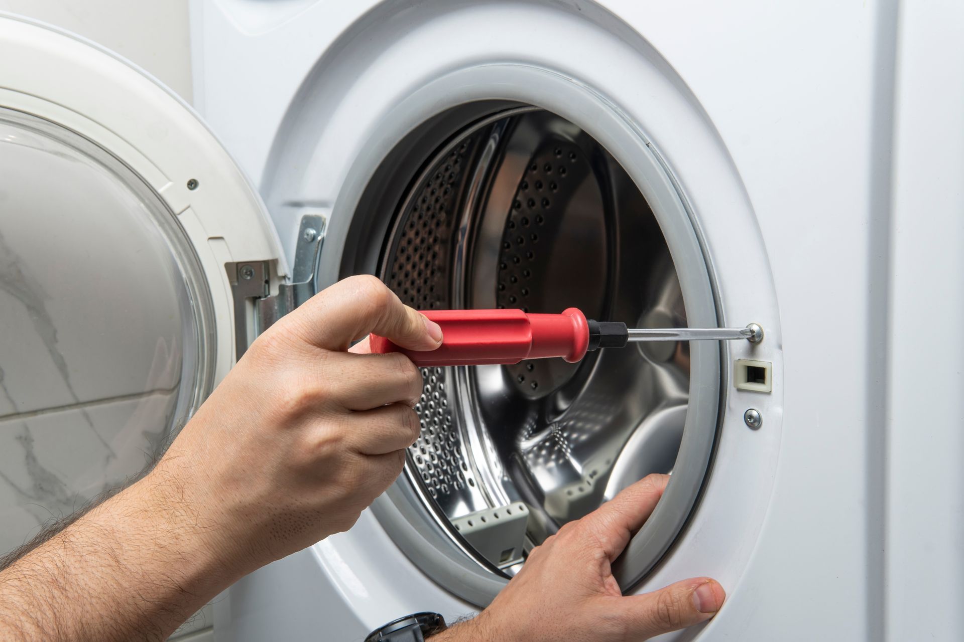 Person fixing a washing machine door with a screwdriver; white appliance, indoor setting.