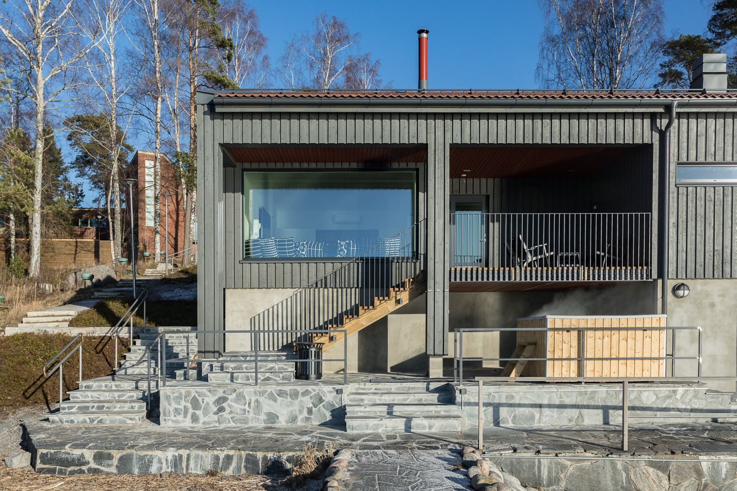 Moderni mökki, jossa on tumma puuverhous, suuri panoraamaikkuna, kiviportaat ja puinen poreallas aurinkoisena päivänä.