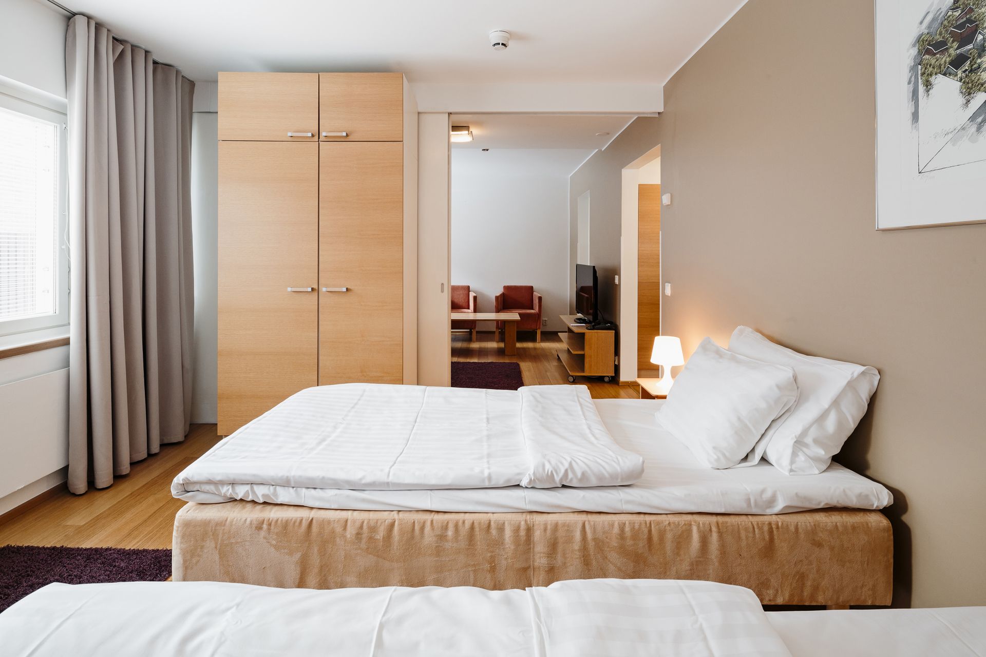 Valoisa hotellihuone, jossa on kaksi sänkyä, puinen vaatekaappi ja näkymä viereiseen olohuoneeseen, jossa on nojatuoleja.