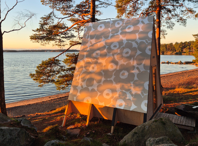 Vaaleasta puusta tehty A-mallinen mökki kuviokatolla sijaitsee meren rannalla mäntyjen keskellä.