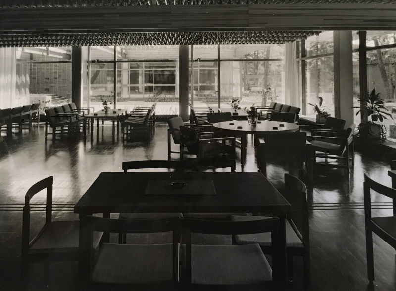 Yksivärinen sisäkuva modernista ruokasalista, jossa on tummat pöydät ja tuolit suurten lasi-ikkunoiden lähellä.