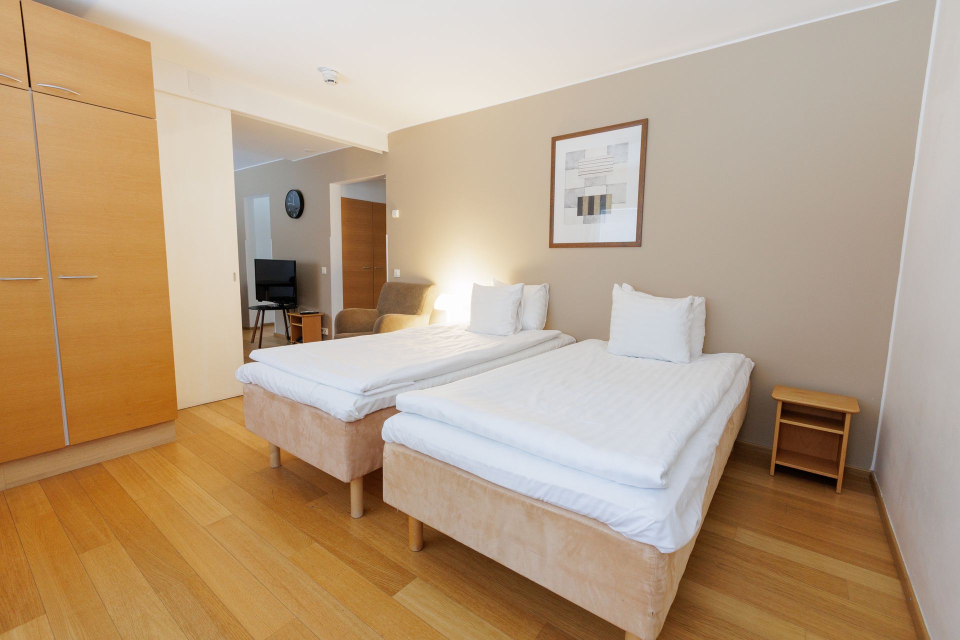 A bedroom with two single beds, white bedding, light-wood flooring, a tall wooden wardrobe, and a small side table.
