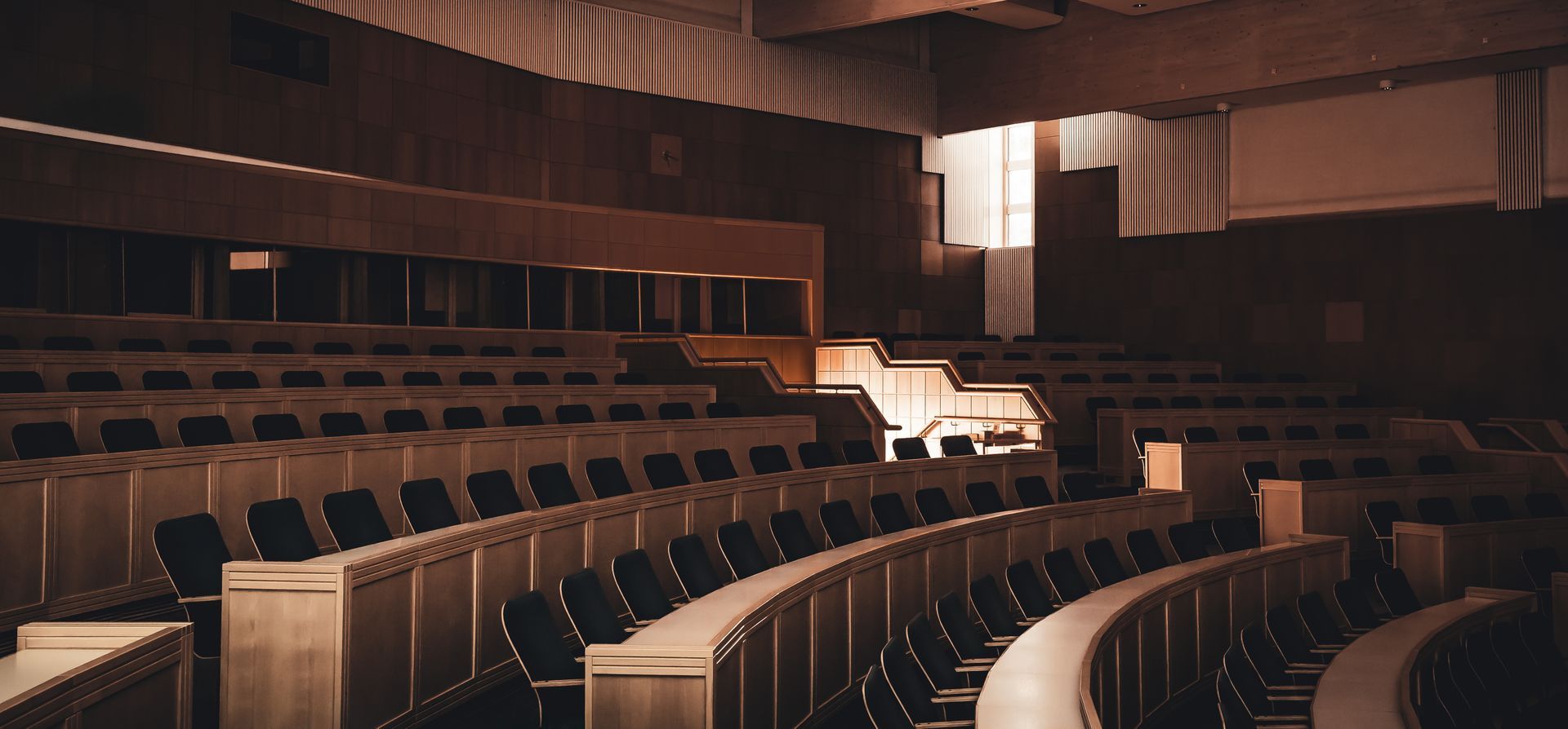 Tyhjä luentosali, jossa on rivit tummia tuoleja puoliympyrän muotoon porrastetusti vaaleilla puupöydillä.