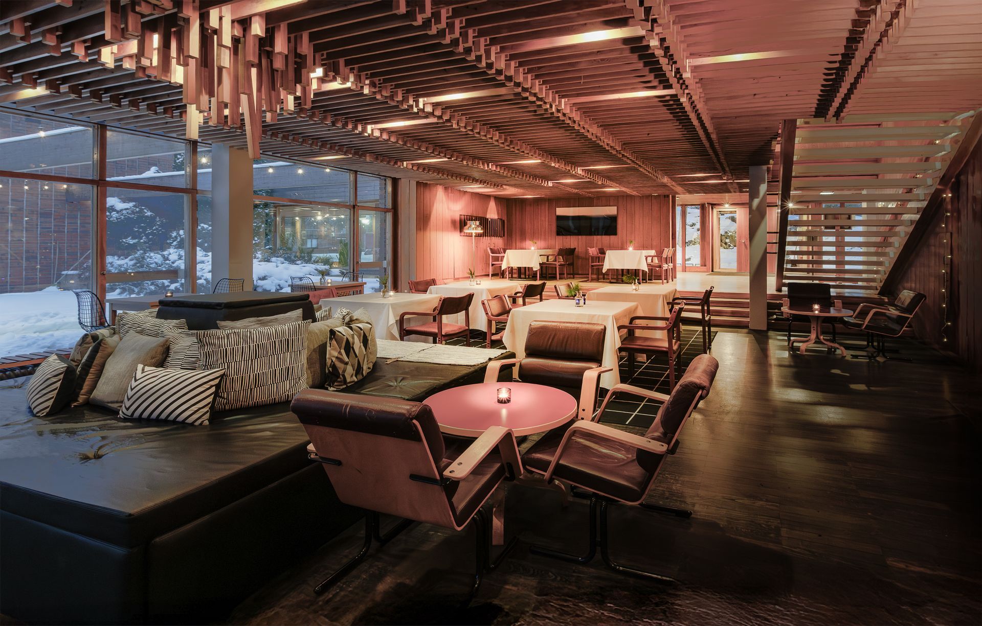 A dimly lit lounge with modern seating, tables, and a distinctive slatted wood ceiling, overlooking a snowy landscape.