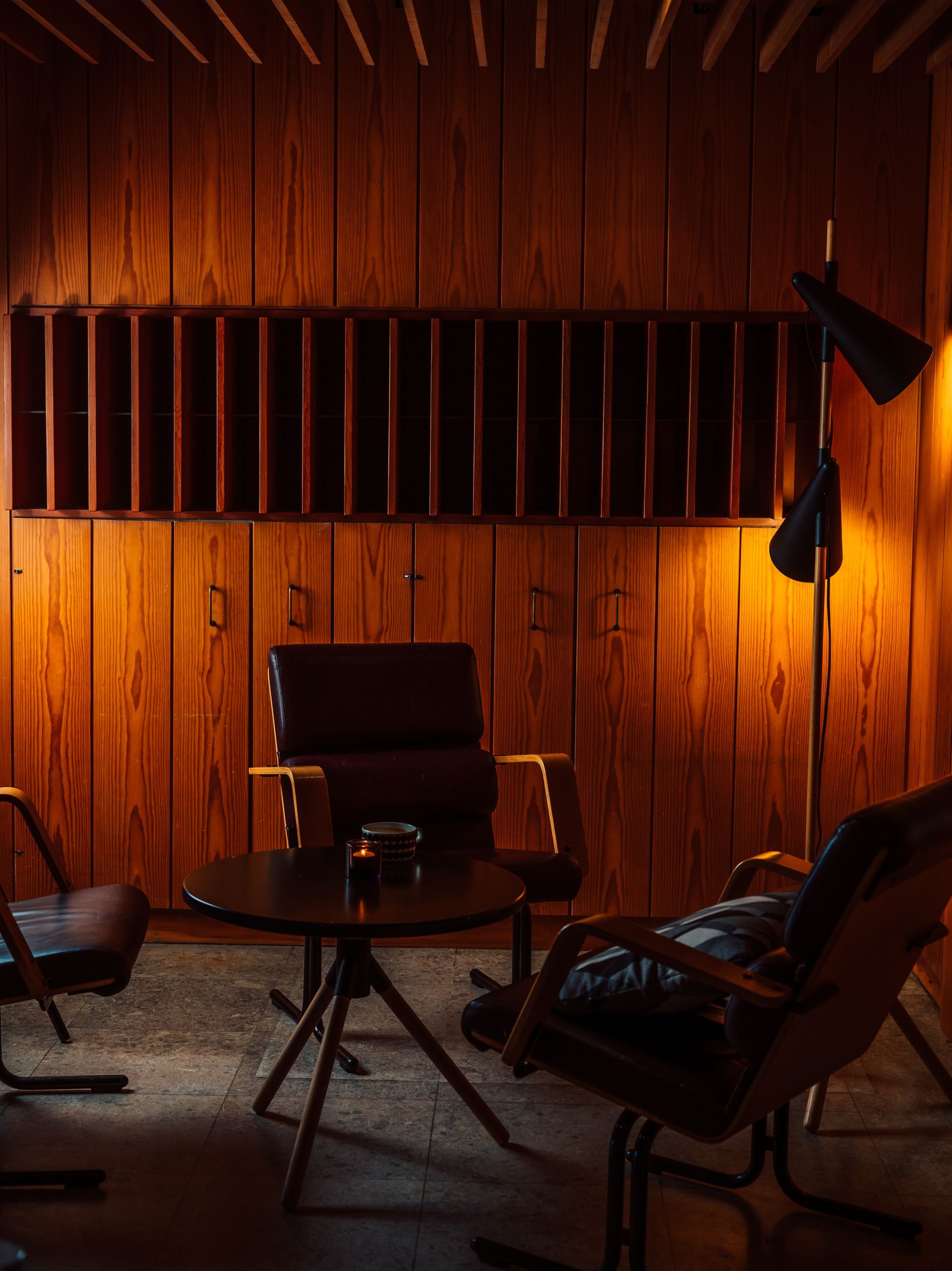 1900-luvun puolivälin moderni oleskelualue, jossa on puupaneeliseinät, pieni pyöreä pöytä ja kolme tummaa nojatuolia, joita valaisee lattiavalaisin.