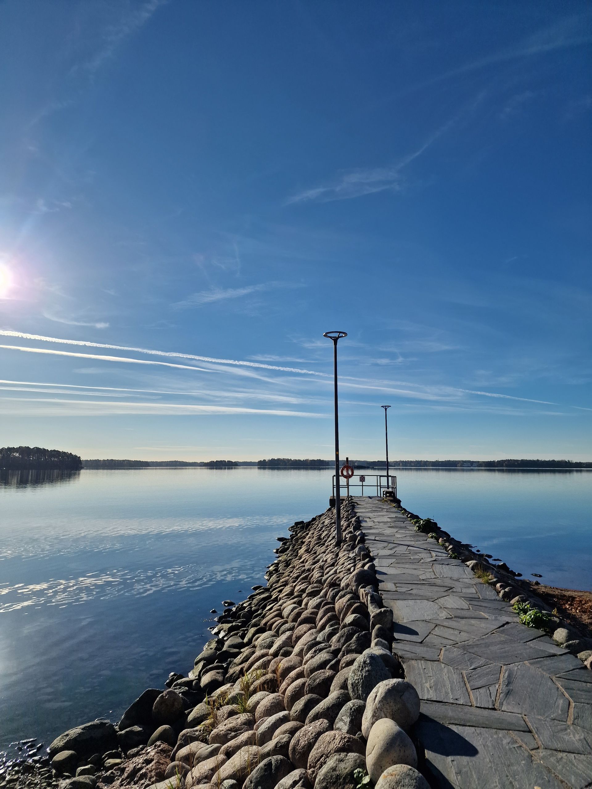 Kivinen laituri ulottuu rauhalliseen, heijastavaan järveen kirkkaan, kirkkaan sinisen taivaan, ohuiden pilvien ja matalalla paistavan auringon alla.