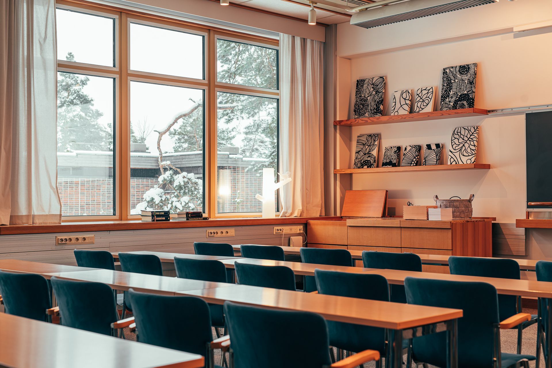 Tyhjä luokkahuone, jossa on rivejä tummansinisiä tuoleja vastakkain puisten pöytien kanssa, suuret ikkunat verhoineen ja hyllyt taideteoksineen.
